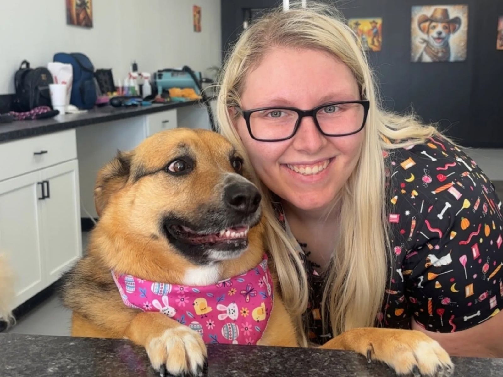 A smiling Dapper Doodle groomer leaning cheek to cheek with a freshly groomed shepherd mix in a pink bandana